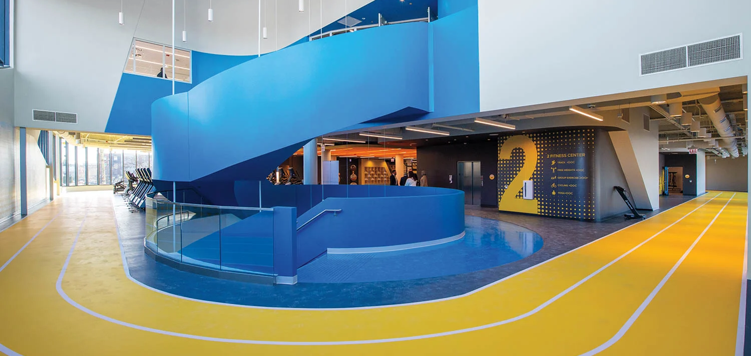 Spacious modern gym interior with a striking blue spiral staircase, yellow running track, and high ceilings.