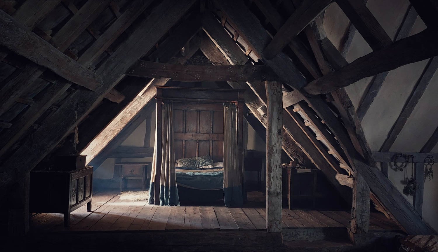 A rustic attic room with wooden beams features a cozy, canopied bed in soft light