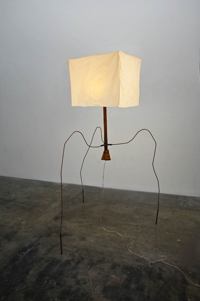 A floor lamp with a rectangular white fabric shade, thin curved metal legs, and a visible electrical cord stands on a concrete floor against a plain wall.