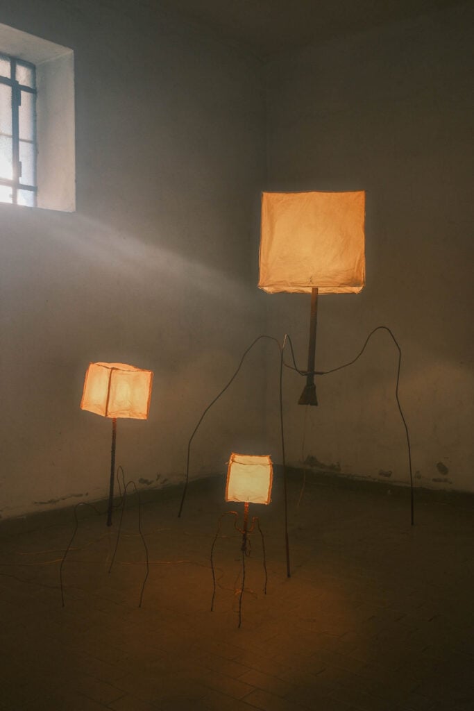 Three box-shaped lamps with glowing shades stand on thin, wire-like legs in a dimly lit, empty room with a barred window.