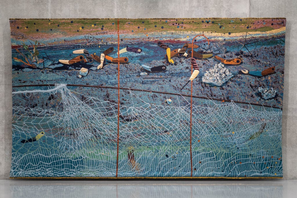 A large triptych painting depicts a shoreline scene with scattered debris, shoes, and fishing nets, set against a textured blue background and concrete wall.