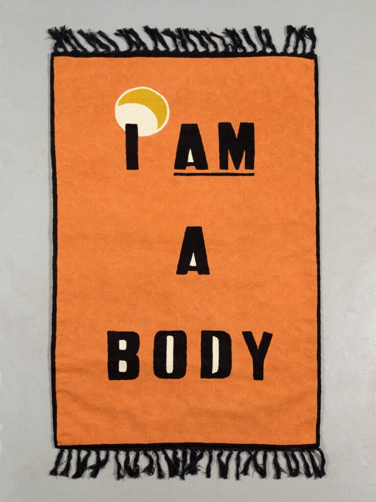 An orange rectangular rug with black fringe displays the words "I AM A BODY" in bold black letters, with a yellow and white circular shape above the letter "I.