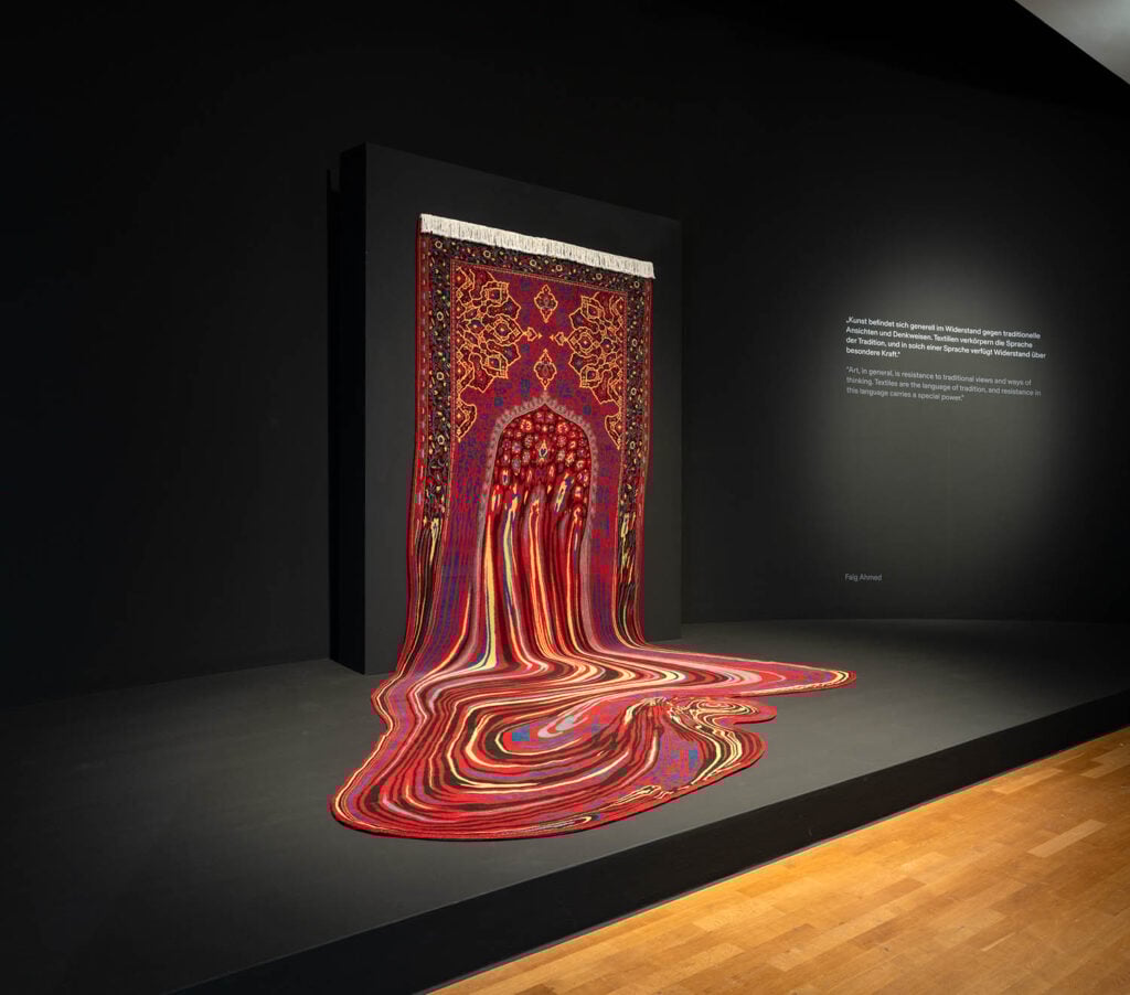 A traditional-style red Persian rug appears to melt and flow onto the floor in a museum exhibition space against a black wall.