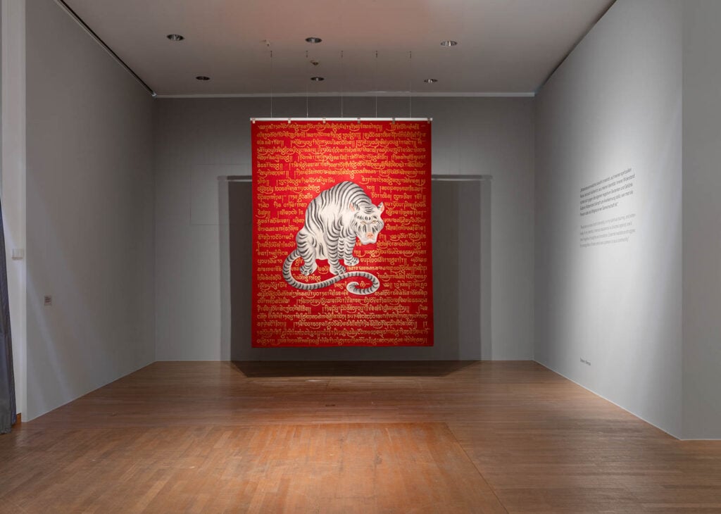 A red textile artwork with a white tiger and script in a gallery space, mounted on a wall with wood flooring and white walls.