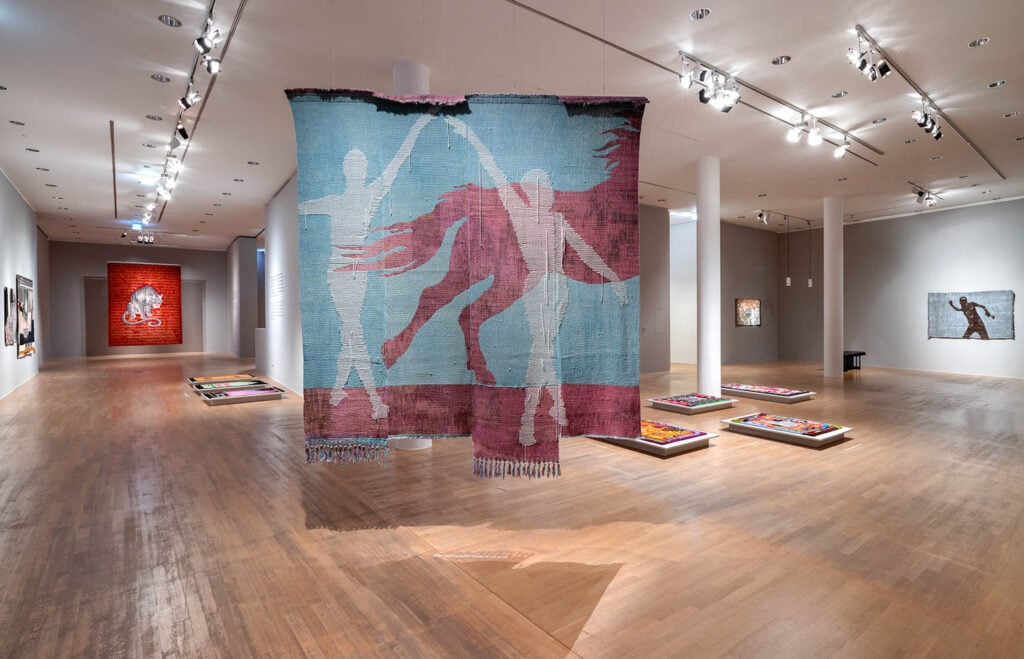 A contemporary art gallery with textile art hanging from the ceiling, featuring silhouetted figures, and other artworks displayed on walls and floors.