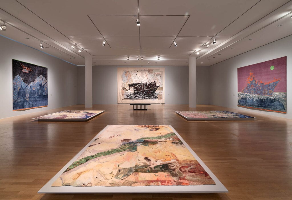 A modern art gallery features large, colorful tapestries on the walls and floor under bright lighting, with a bench centered against the far wall.