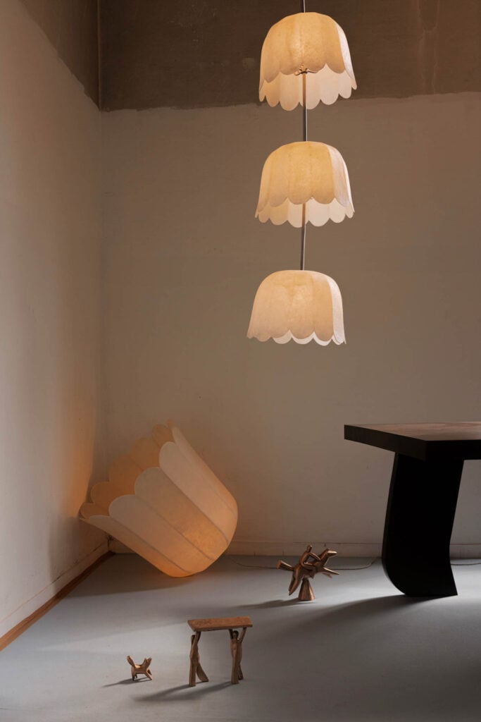 Three layered pendant lights with scalloped edges hang from the ceiling; a large matching lamp and small wooden sculptures are on the floor near a dark table in a minimalist room.