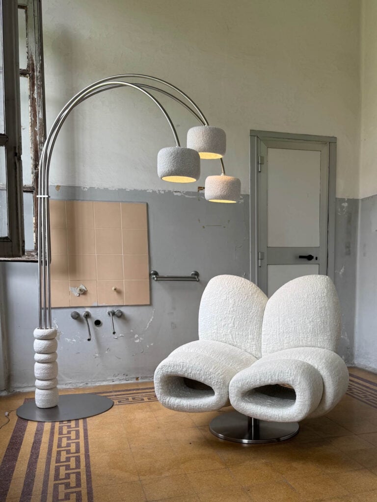 A modern, white textured lounge chair and a three-headed floor lamp are placed in a sparse room with tiled floors, an old sink area, and a gray door.
