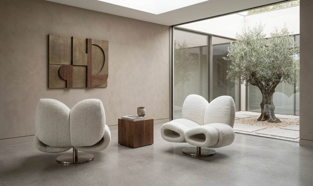 Two modern white chairs and a wooden side table in a minimalist room with abstract wall art and large windows overlooking a tree in an outdoor patio.