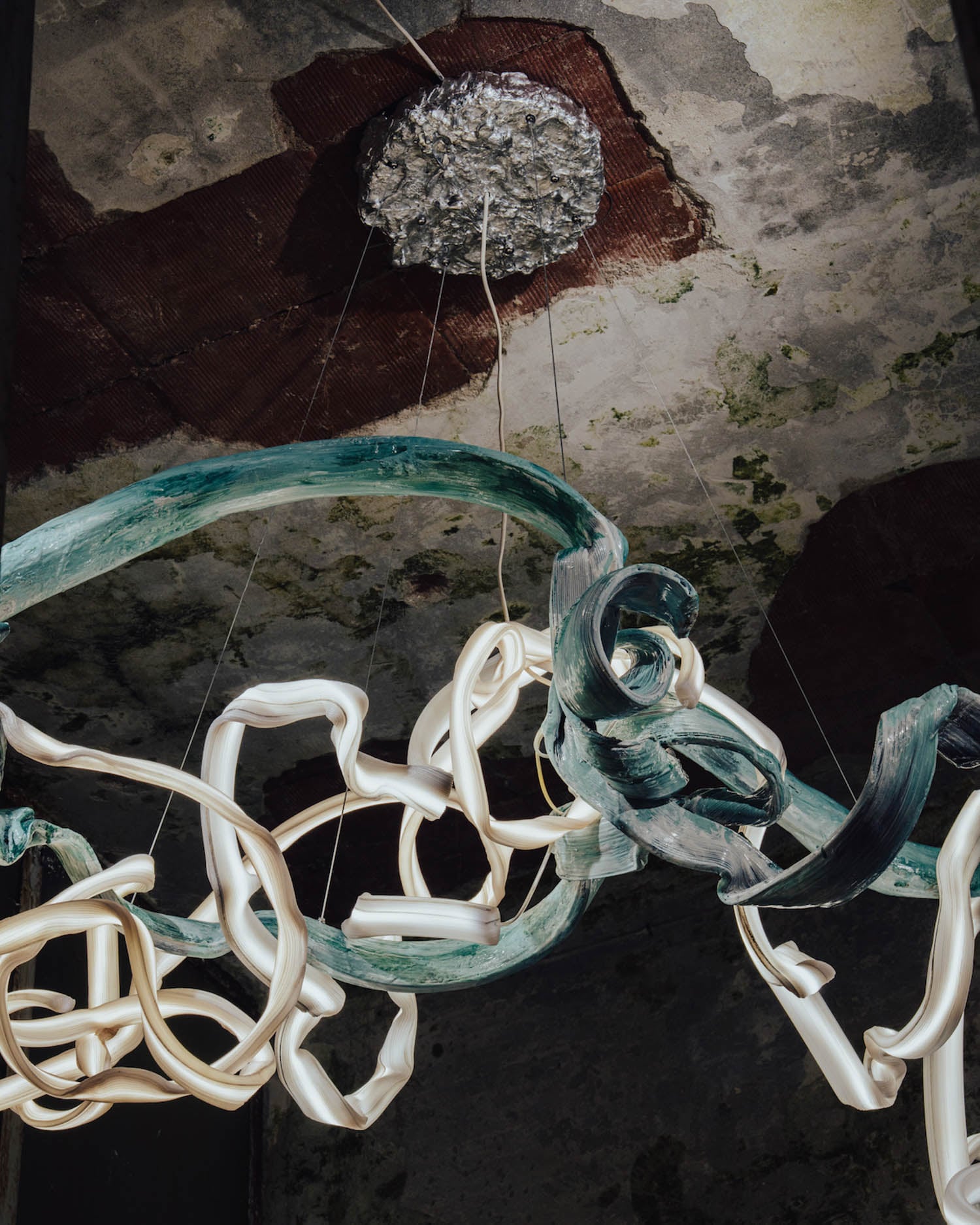 Curved white and green sculptural forms hang from the ceiling by wires, beneath a textured, weathered ceiling with a metallic circular fixture.