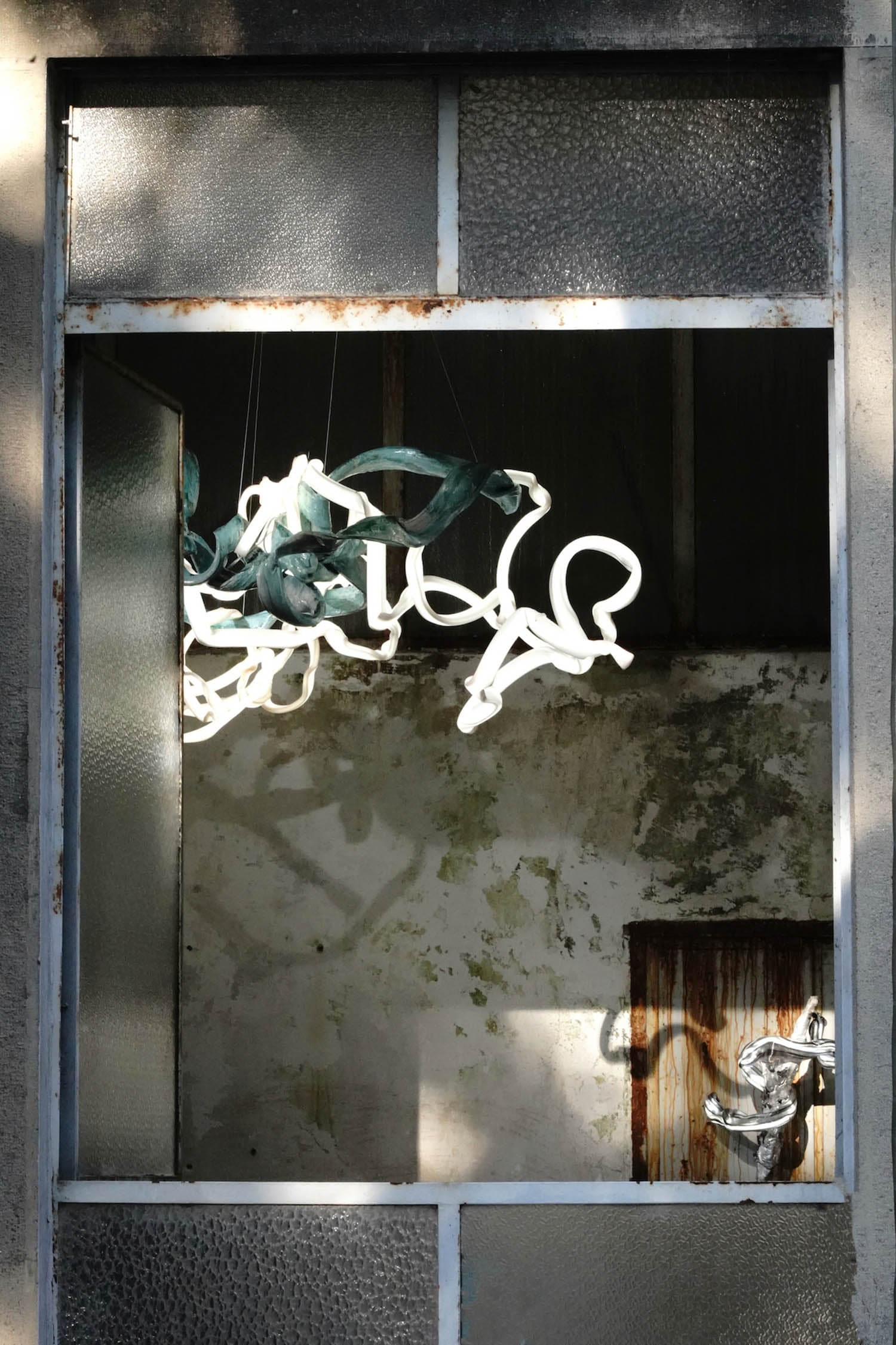 A window with no glass reveals an interior with peeling walls and sculptural, twisted white and green light fixtures hanging from the ceiling.