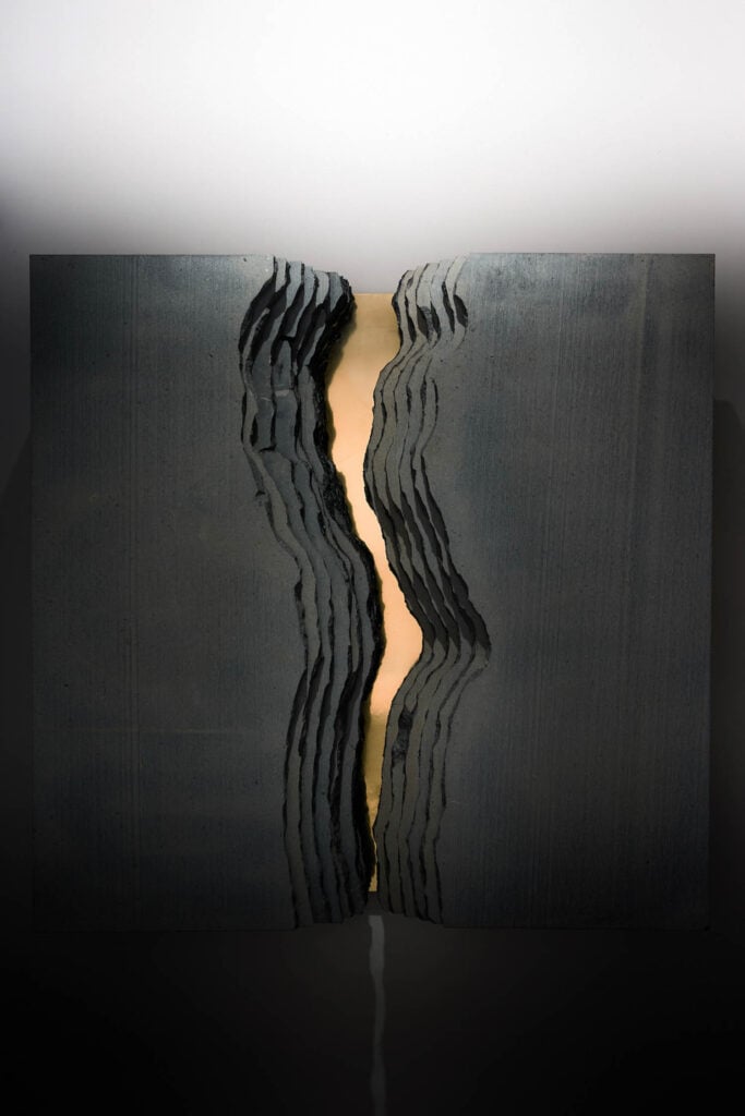 A dark, square sculptural panel with a jagged vertical split in the center, revealing a glowing light from within the crevice.