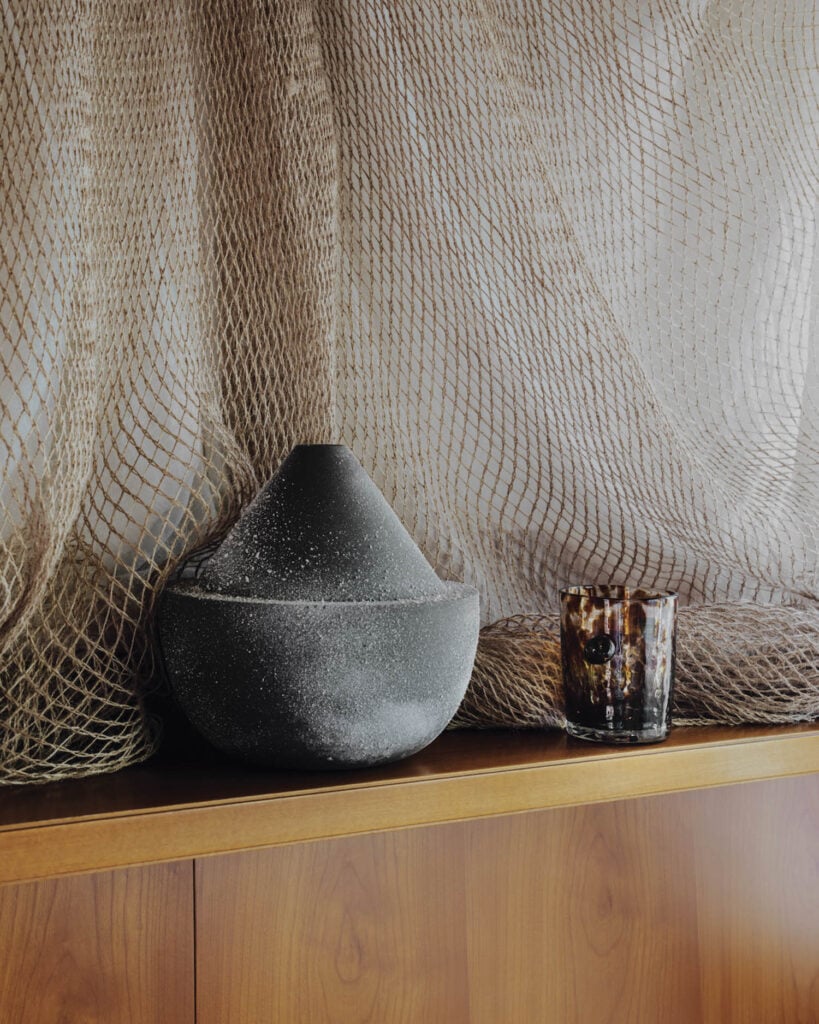 A black textured vase and a small glass cup sit on a wooden shelf in front of sheer, netted curtains.