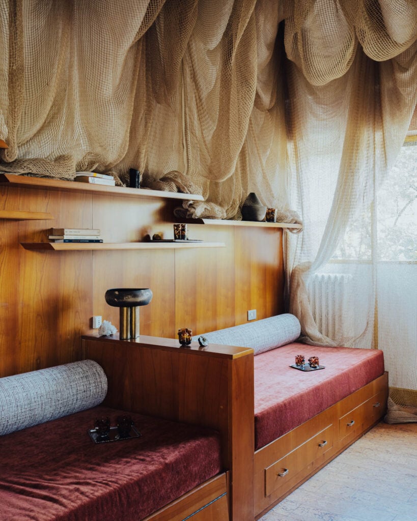 Room with two wooden daybeds with red cushions, shelves with decor items, and fabric netting draped across the ceiling and window. Natural light enters from the right.