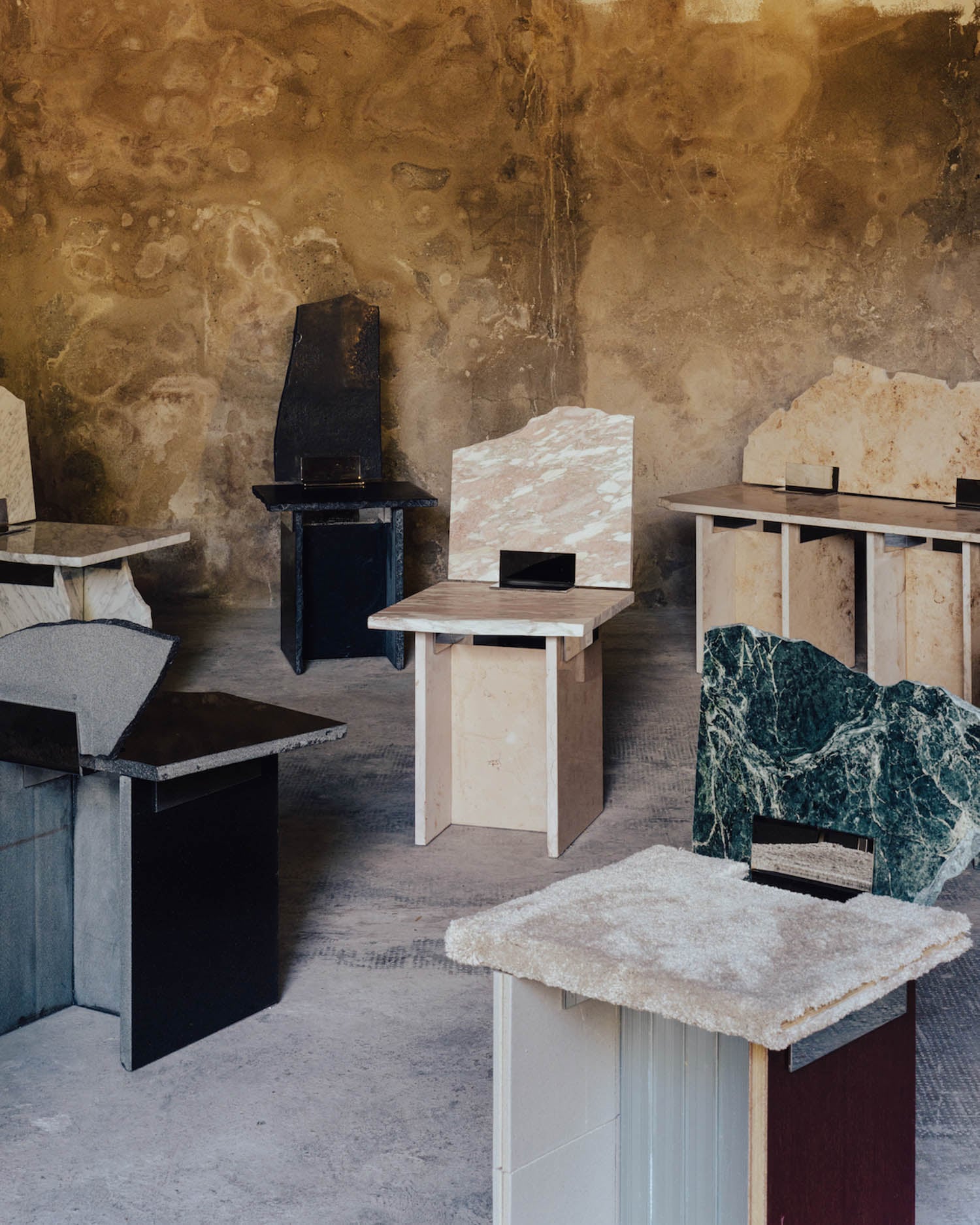 Several uniquely shaped stone chairs with geometric designs are arranged in a rustic room with textured, weathered walls.