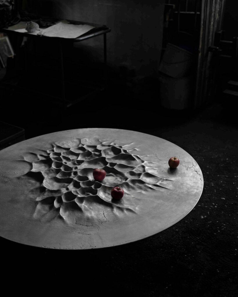 A round, textured metal table with an uneven, cratered surface holds three red apples in a dimly lit industrial space.