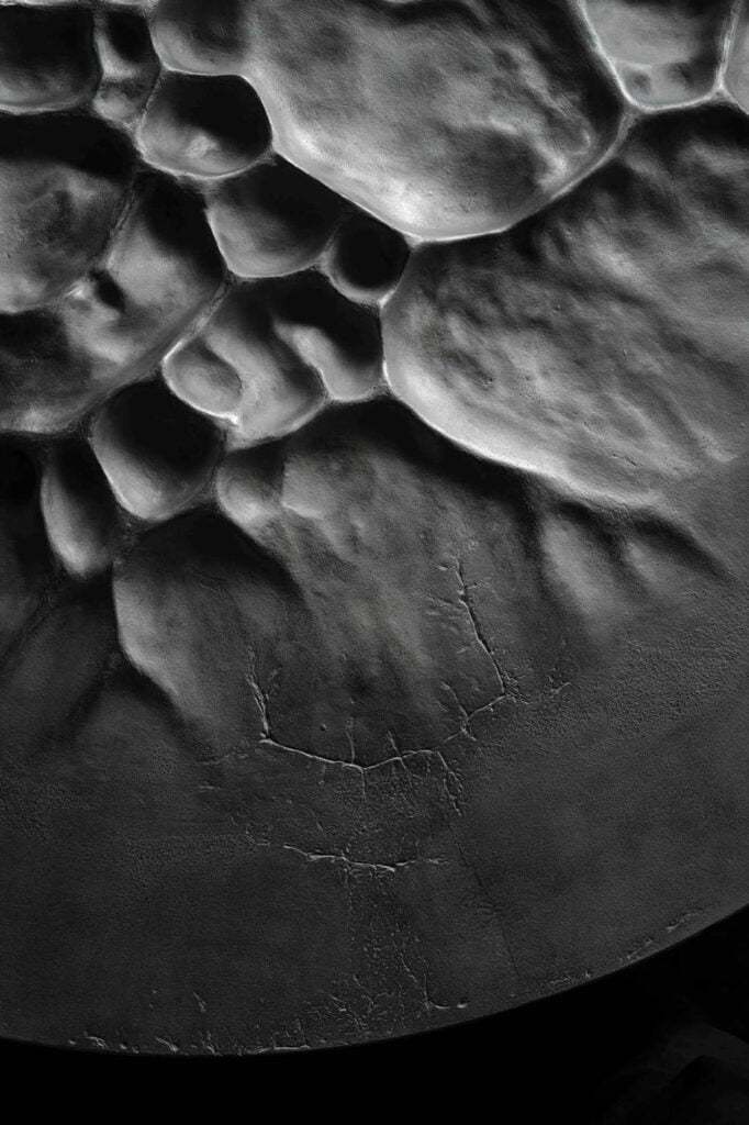 A close-up, black and white image of a textured, uneven surface with cracked and raised areas, resembling geological formations or abstract patterns.