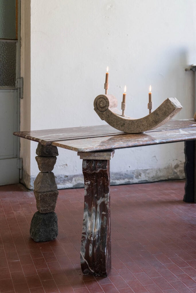 A rustic table with mismatched stone and wood legs stands on a red tile floor, topped with a three-candle stone holder, each candle lit.