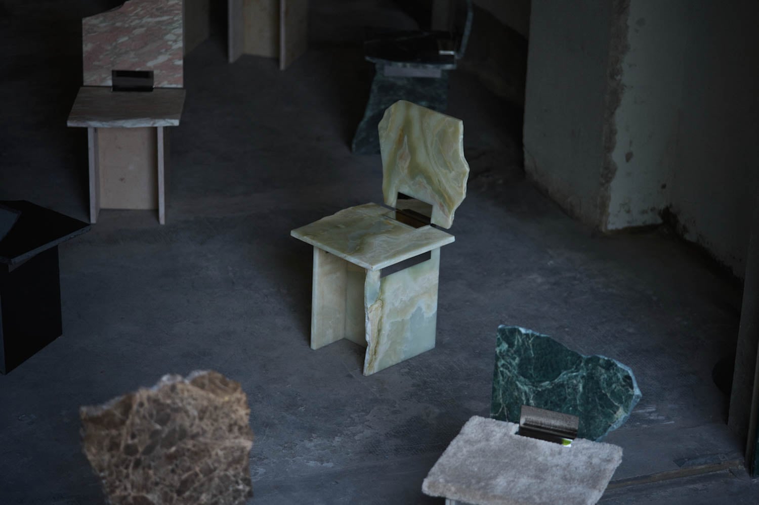 Several uniquely shaped stone chairs with varying colors and textures are arranged on a dark concrete floor in a dimly lit room.