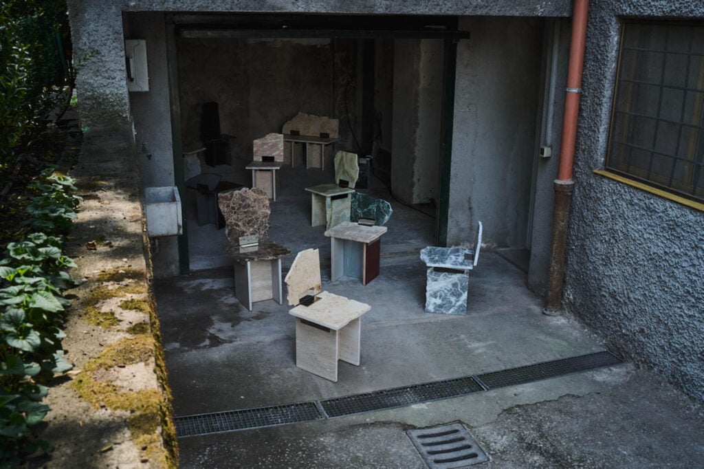 Several uniquely shaped chairs made of stone and other materials are arranged in an open garage with concrete walls and floor.
