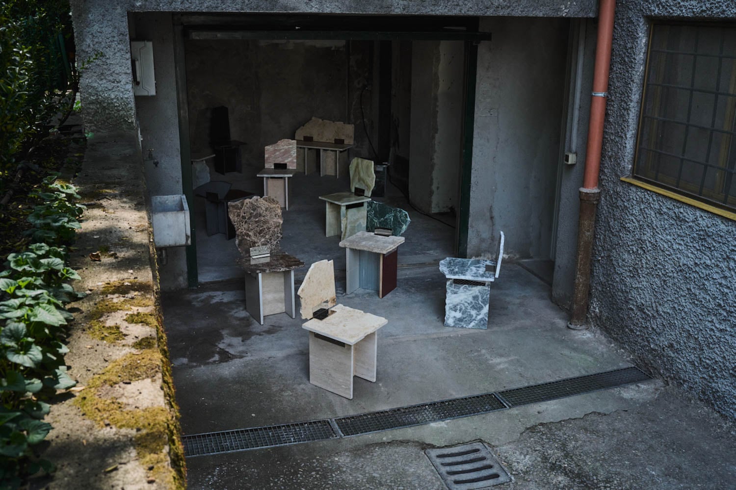 Several uniquely shaped chairs made of stone and other materials are arranged in an open garage with concrete walls and floor.