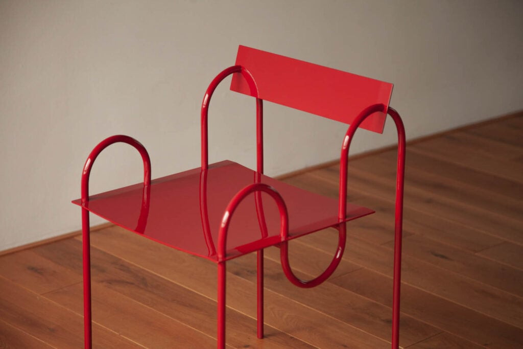 A bright red, modern metal chair with a flat square seat, curved armrests, and a straight backrest, placed on a wooden floor against a light wall.