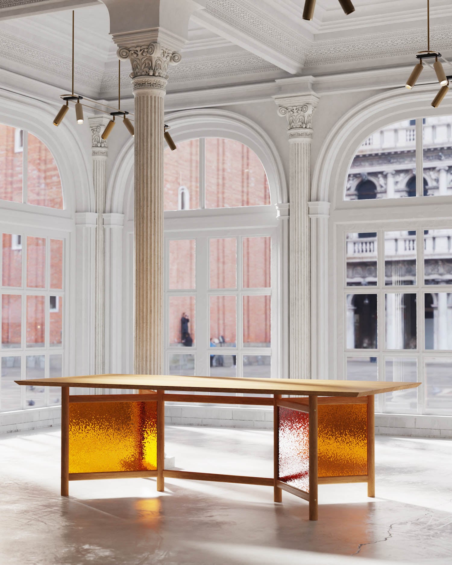 A modern desk with amber-colored textured glass panels stands in a bright room with tall arched windows, ornate columns, and a view of historic buildings outside.