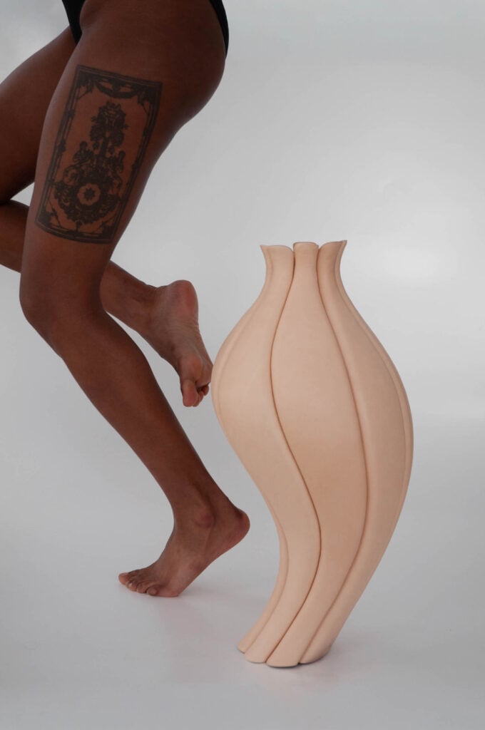 A person with a large thigh tattoo poses on tiptoe next to a sculptural, beige vase with curved, petal-like segments against a plain background.