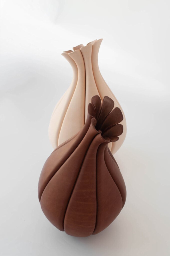 Two sculptural vases with smooth, curving segments; one is beige with a narrow opening, and the other is brown with a flared, petal-like rim. Both are set against a plain white background.
