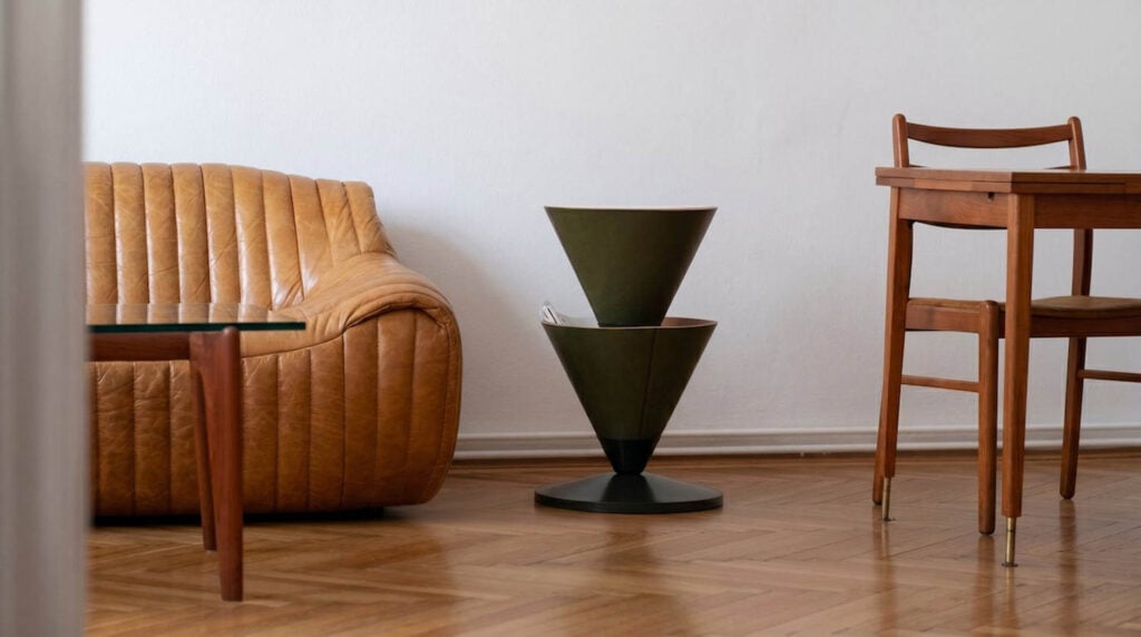 A tan leather sofa, a wooden dining table with chairs, and a large modern planter sit on a wooden floor against a plain white wall.