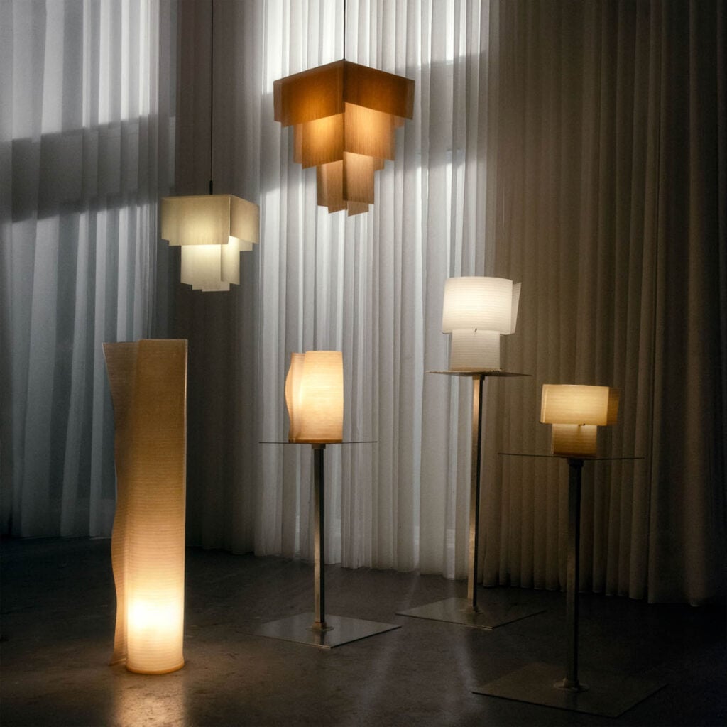 A group of five modern lamps with geometric, translucent shades are arranged in a dimly lit room with sheer curtains.
