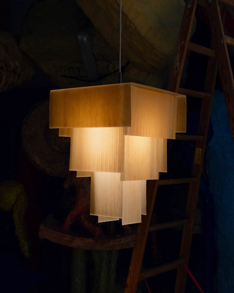 A geometric pendant lamp with layered rectangular panels emits a warm light, hanging in front of a wooden ladder in a dimly lit room.