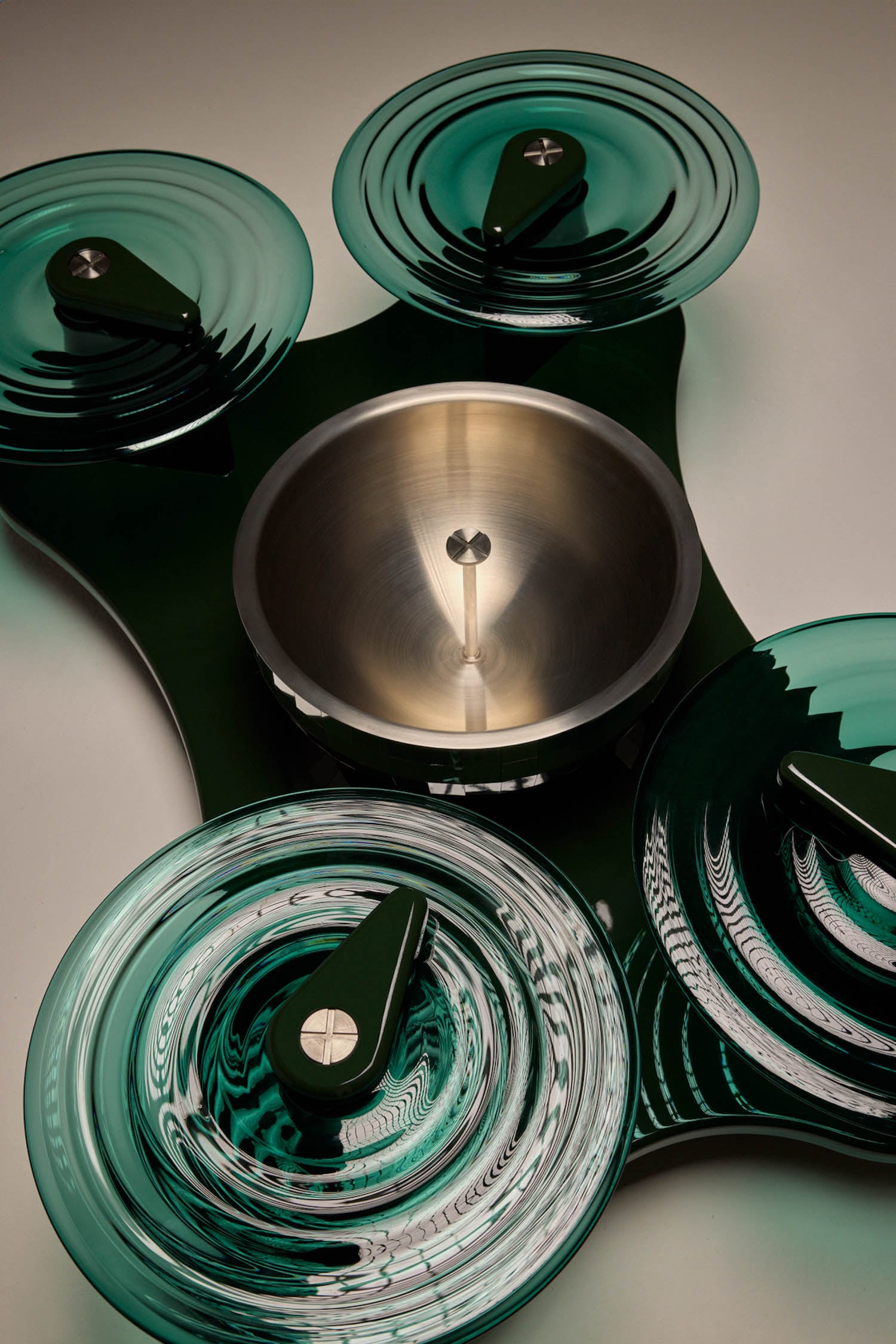 A metal bowl is centered on a dark base, surrounded by four green glass dishes with matching green handles, arranged symmetrically.