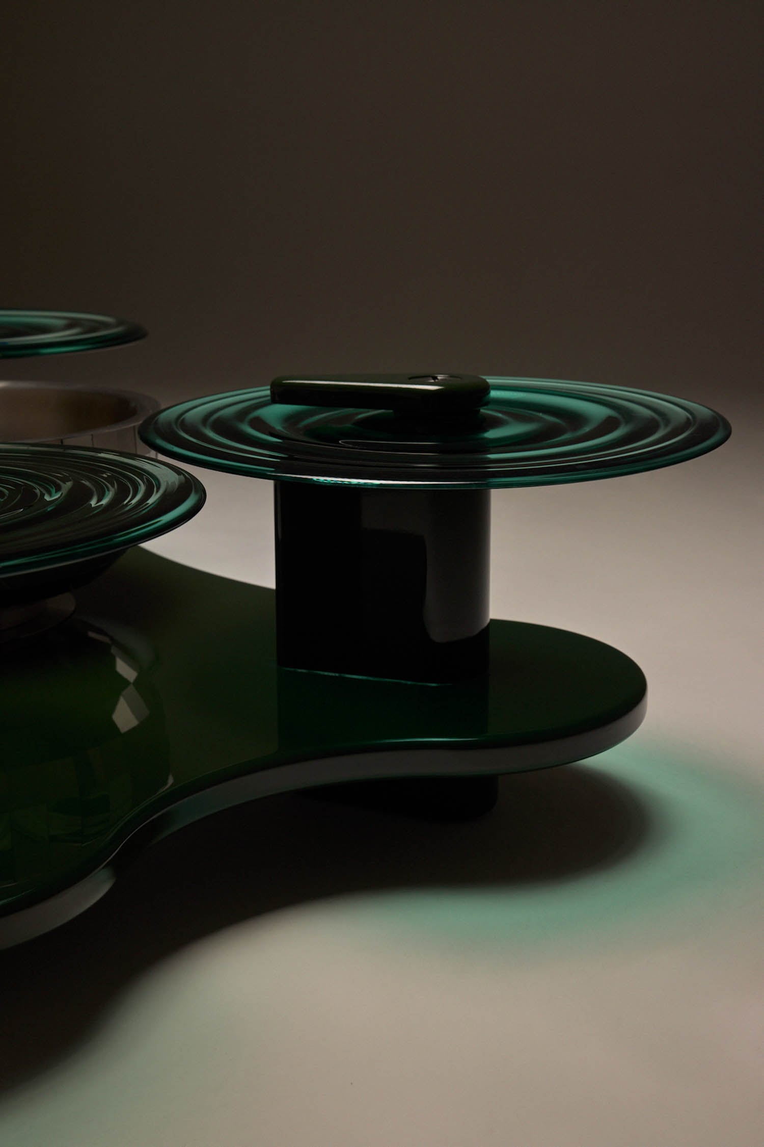 A sculptural object with glossy dark green surfaces and concentric circular patterns, displayed on a flat base against a neutral background.