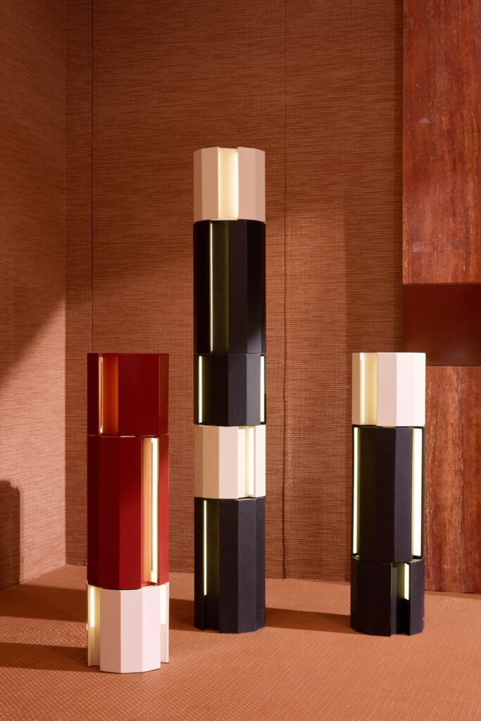 Three geometric, column-like floor lamps in red, black, and cream colors stand on a textured brown floor against a matching wall, with warm lighting highlighting their shapes.