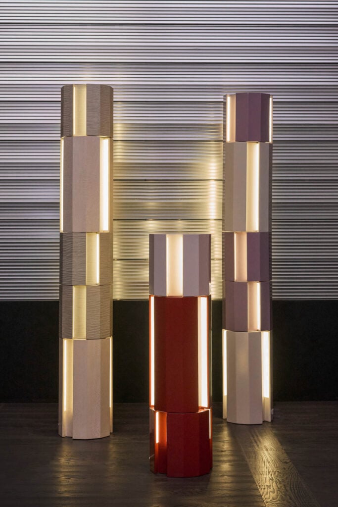 Three geometric column lamps with alternating colored segments emit soft light, standing on a dark surface in front of a ribbed metallic wall.