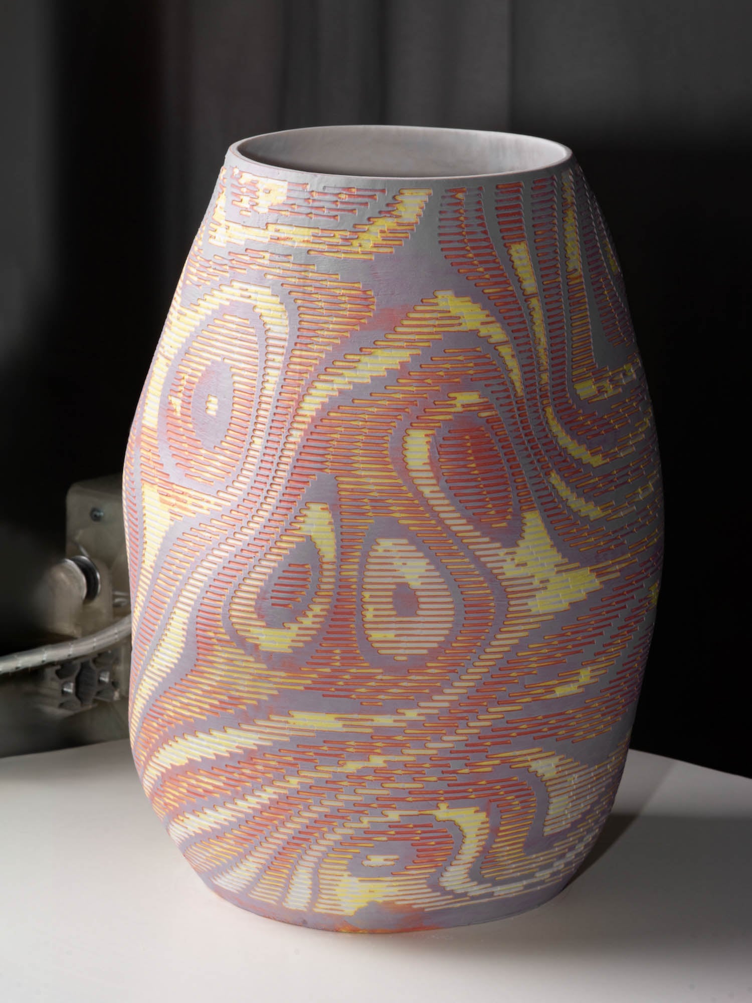 A large ceramic vase with a matte surface, featuring intricate, wavy patterns in shades of yellow, orange, and lavender, placed on a white surface against a dark background.