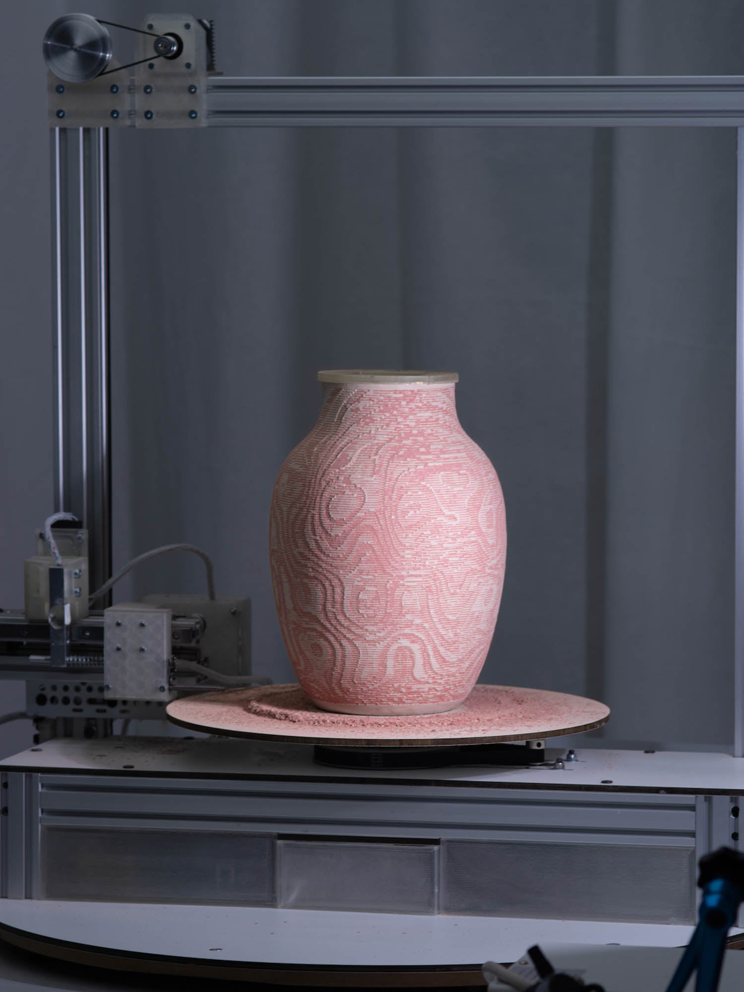A pink ceramic vase with textured, swirling patterns sits on a rotating platform inside a metal-framed machine.