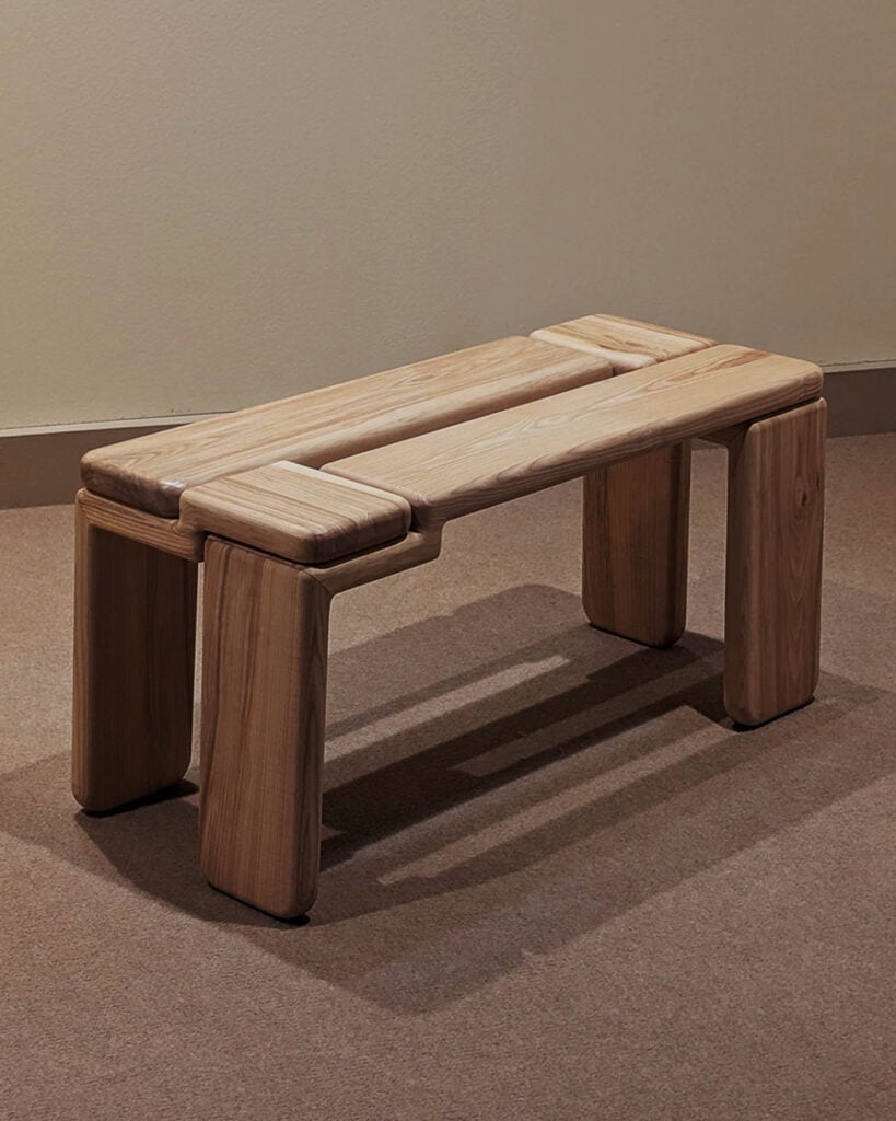 A wooden bench with a minimalist design, featuring four sturdy legs and a smooth, rectangular seat, placed on a carpeted floor against a plain wall.