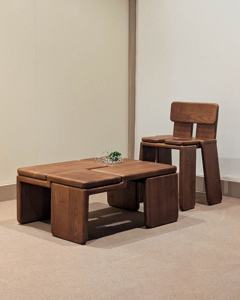 A wooden chair and matching low table with curved edges are placed on a beige carpet against a plain, light-colored wall. A small sprig of greenery rests on the table.