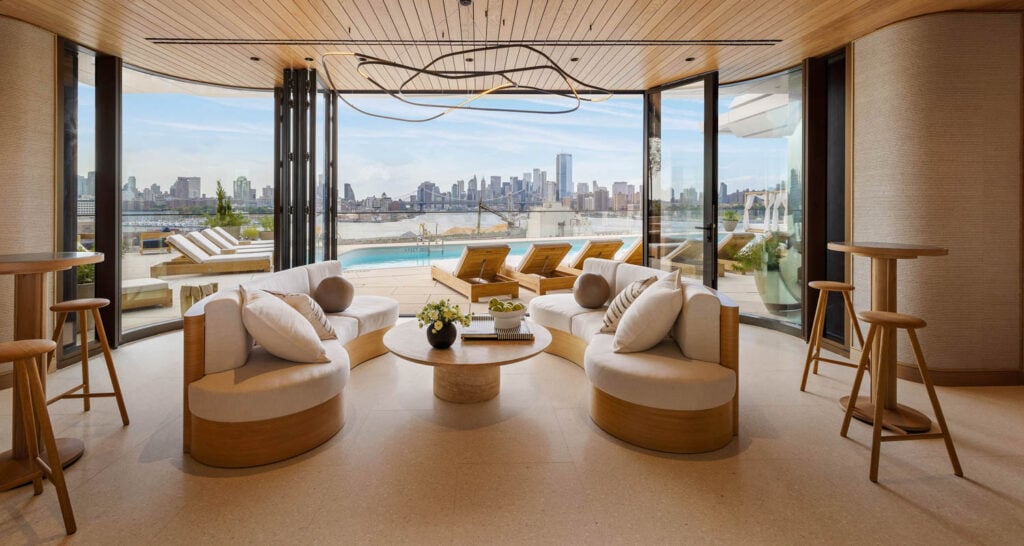 Modern lounge area with round gray chairs, wooden tables, and large windows overlooking a pool and city skyline in the background—a stylish urban ward for relaxation.