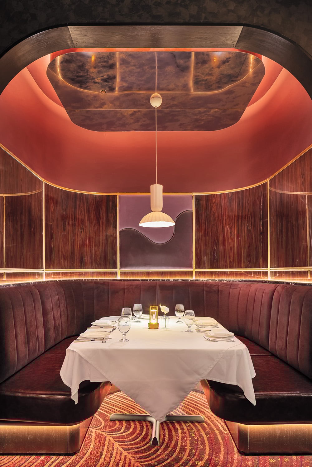 A round booth with dark red velvet seating surrounds a white tablecloth dining table set for six, under warm lighting and a modern pendant lamp in a wood-paneled restaurant alcove.