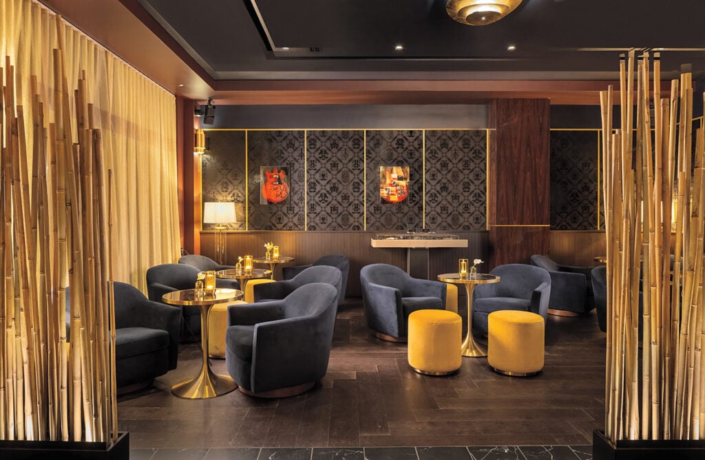 Modern lounge with navy blue chairs, round golden tables, and yellow ottomans, separated by tall bamboo decor; dim lighting and decorative wall art create a cozy atmosphere.
