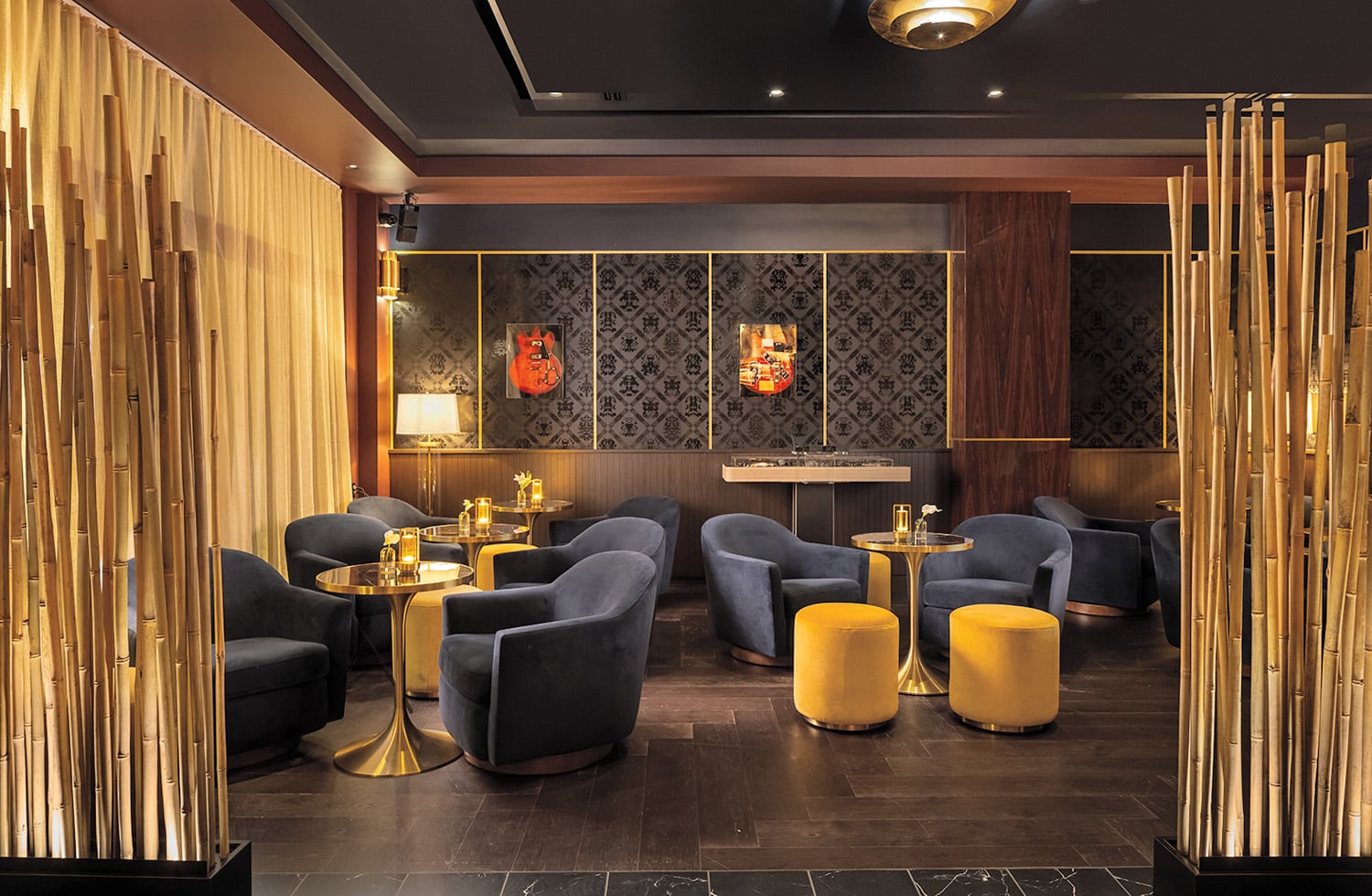 Modern lounge with navy blue chairs, round golden tables, and yellow ottomans, separated by tall bamboo decor; dim lighting and decorative wall art create a cozy atmosphere.