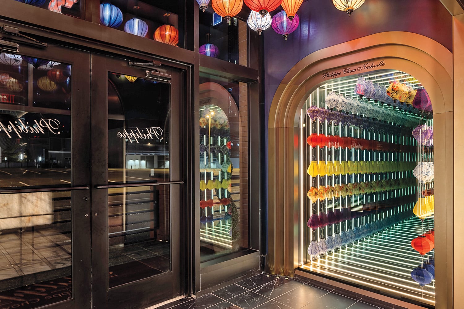 Colorful feather dusters are arranged in rows inside a mirrored display case near a glass entryway, creating an illusion of infinite reflection. Lanterns hang from the ceiling above.