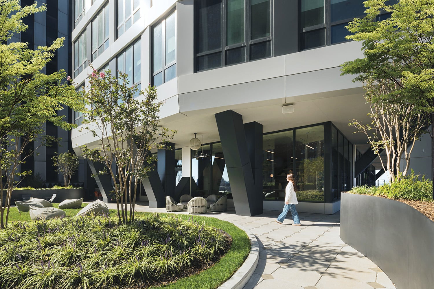 A person walks on a curved sidewalk next to a modern building with large windows and landscaped greenery in the foreground.