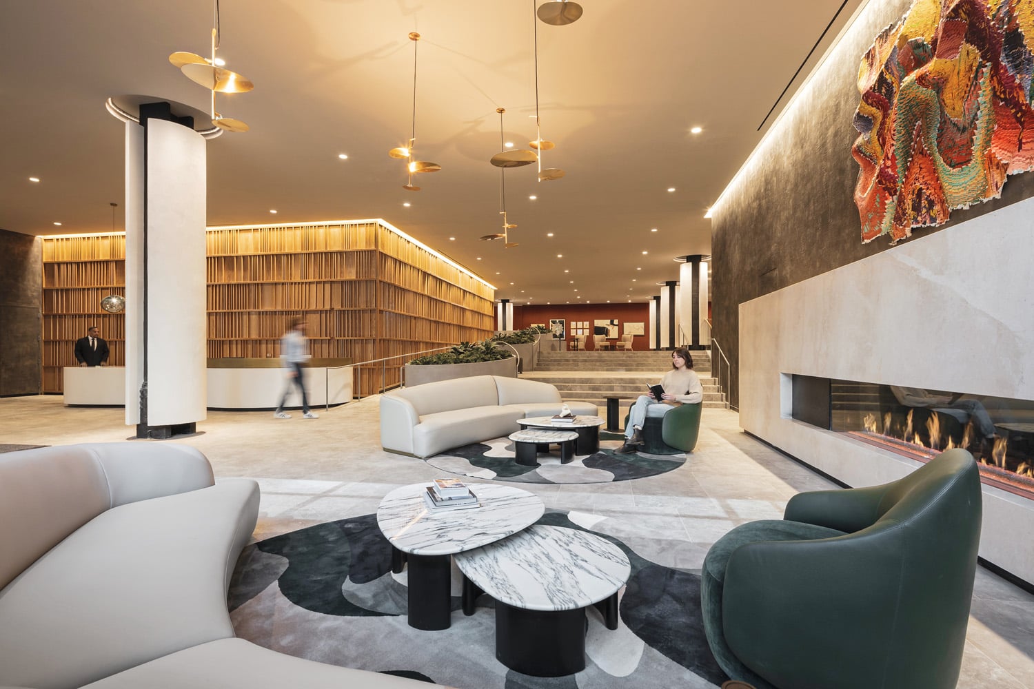 Modern hotel lobby with stylish furniture, marble tables, a built-in fireplace, wall art, and a person seated; another person walks near the reception desk in the background.