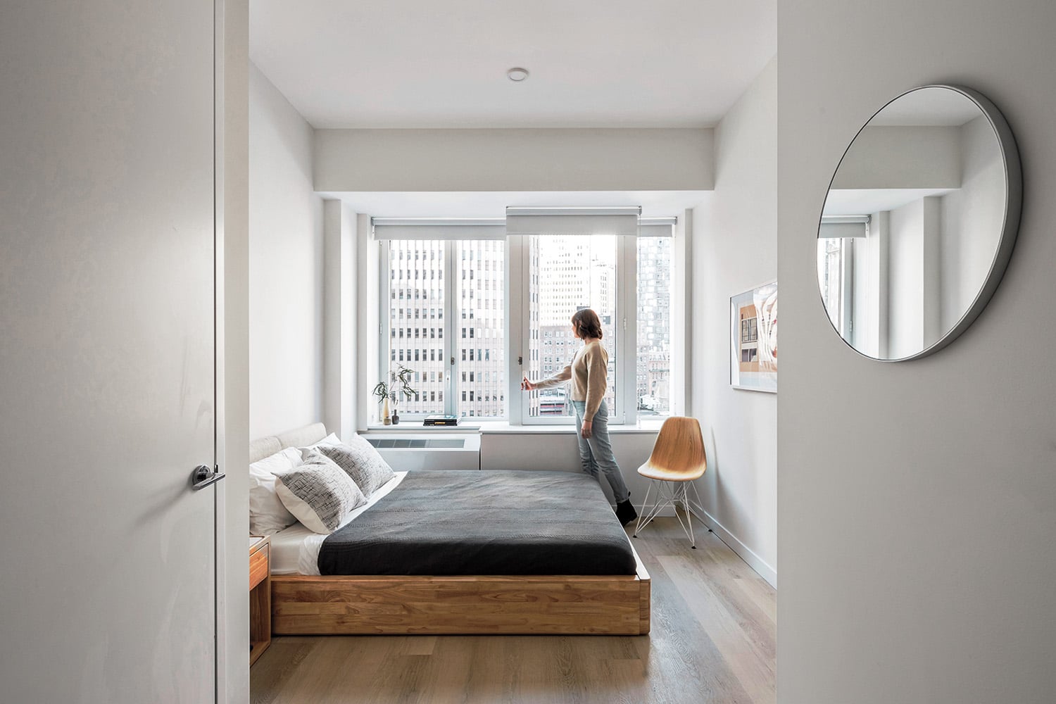 Person standing by large windows in a bright, minimalist bedroom with a wooden bed, white walls, and modern decor.