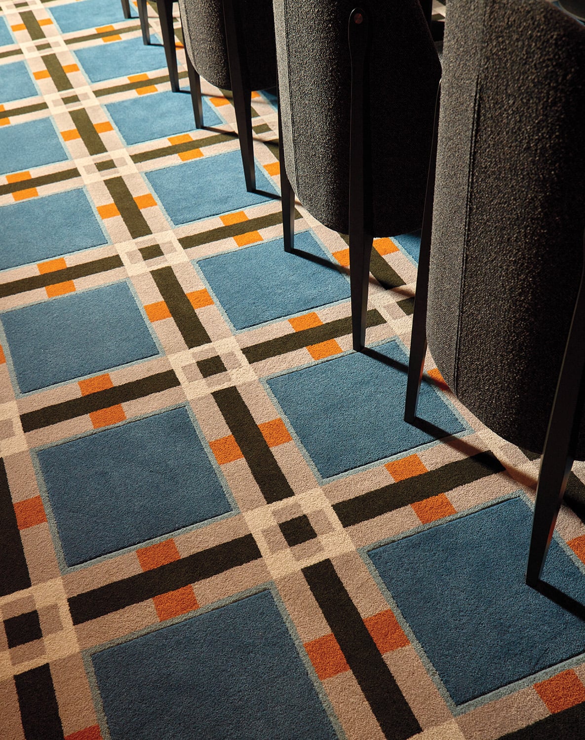 Rows of dark chairs sit on a carpet with a geometric pattern featuring blue squares, white and dark lines, and orange accents.