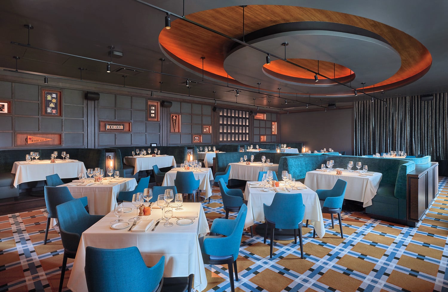 Modern restaurant dining area with blue chairs, white tablecloths, geometric carpet, and decorative ceiling lighting. Tables are set for guests, and framed art hangs on the walls.
